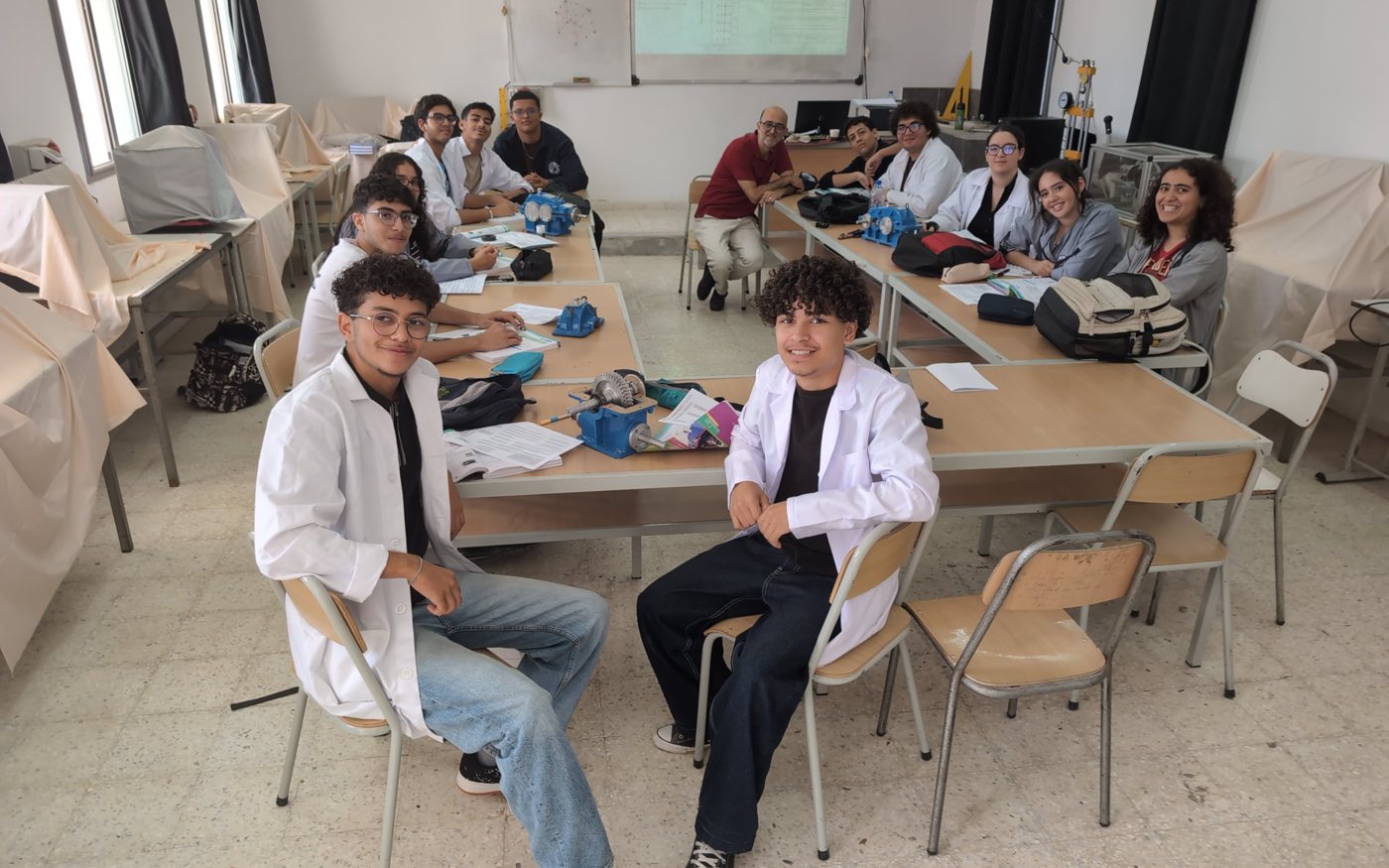 Students in lab coats around tables with gear reducers, their teacher at the end of the table